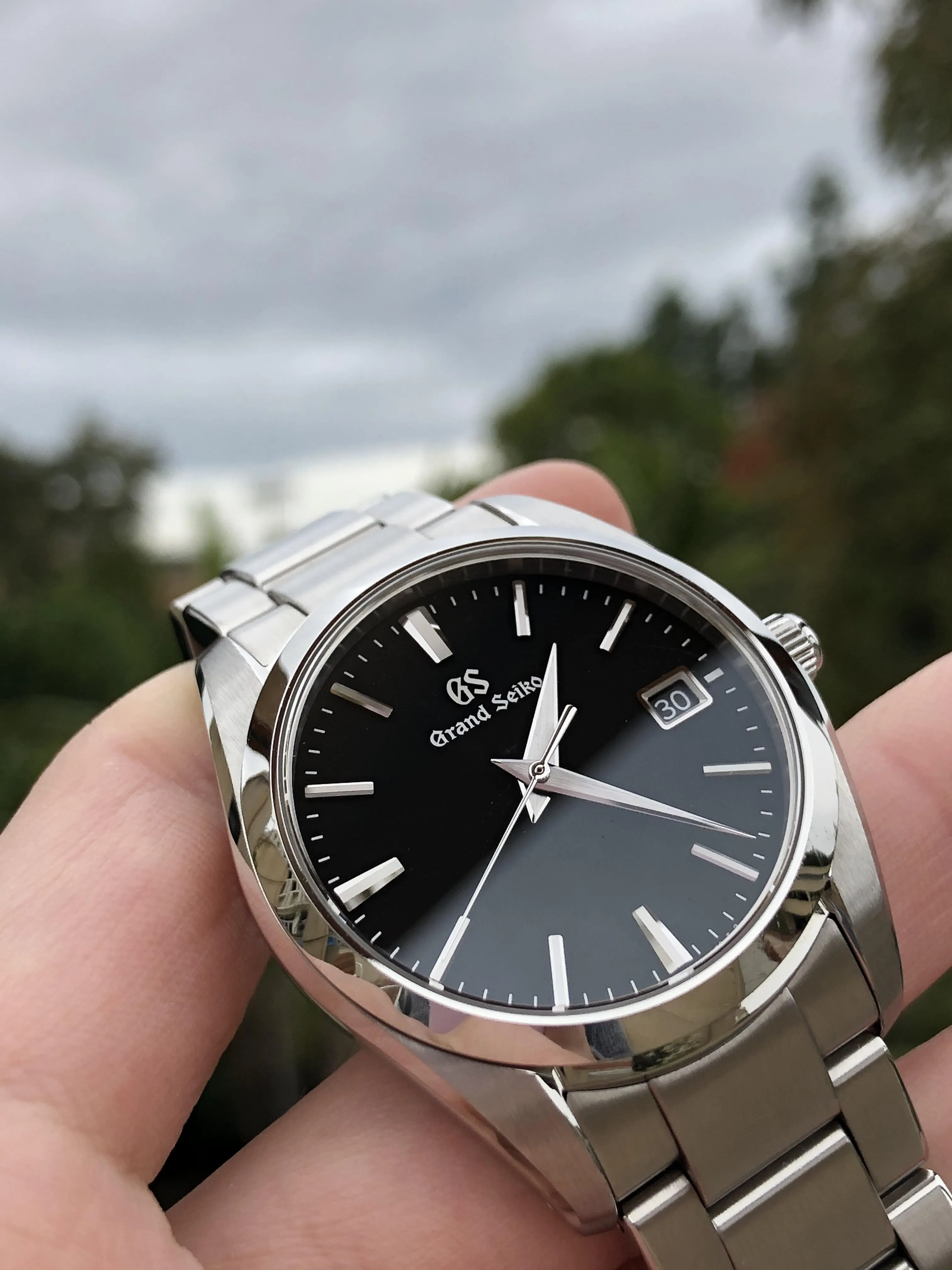 Grand Seiko SBGX261 black dial held face-on outdoors, overcast sky background